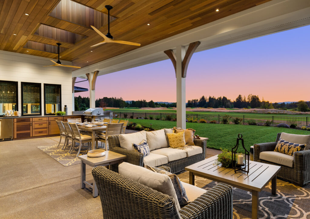 Exterieur eines Hauses bei Sonnenuntergang: überdachte Außenterrasse mit Küche, Grill, Esstisch und Sitzbereich mit Blick auf Wiese und Bäume.