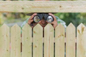 Neugieriger Nachbar steht hinter einem Zaun und schaut mit Fernglas rüber zum Nachbarn.
