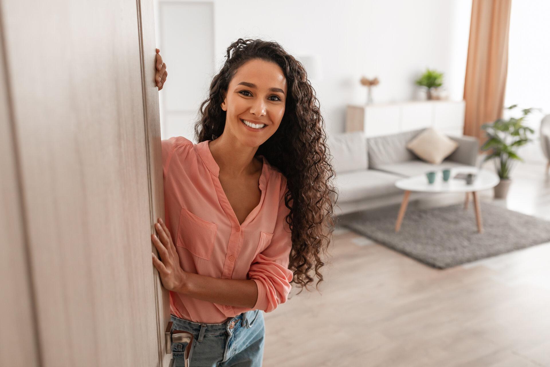Porträt einer fröhlichen jungen Frau, die in der Tür eines neuen, modernen Hauses steht und einen Besucher empfängt, der sie einlädt, ihre stilvolle Wohnung zu betreten